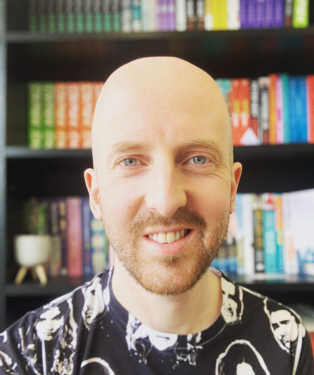 Bert Headshot smiling in front of a colourful bookshelf