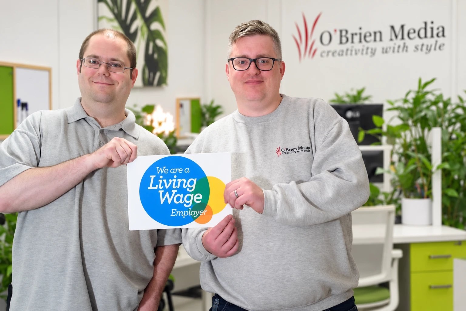 Two O’Brien Media team members holding a “We are a Living Wage Employer” sign in the office with a link to O’Brien Media is accredited Living Wage Employer