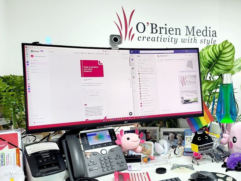 The O’Brien Media office desk with a wide monitor, desk phone and the company logo on the wall behind with a link to Contact
