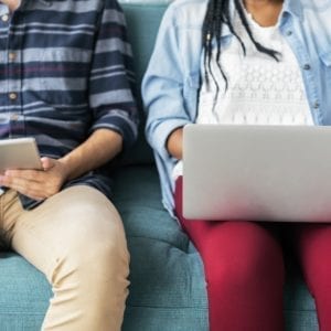 two persons sitting on a sofa, one checking his tablet and another person checking her laptop
