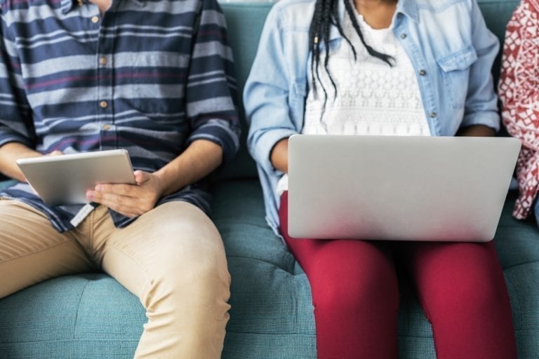 two persons sitting on a sofa, one checking his tablet and another person checking her laptop