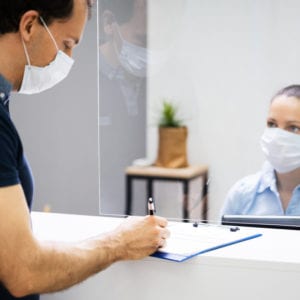 Client Signing Paper At Reception Desk In Face Mask