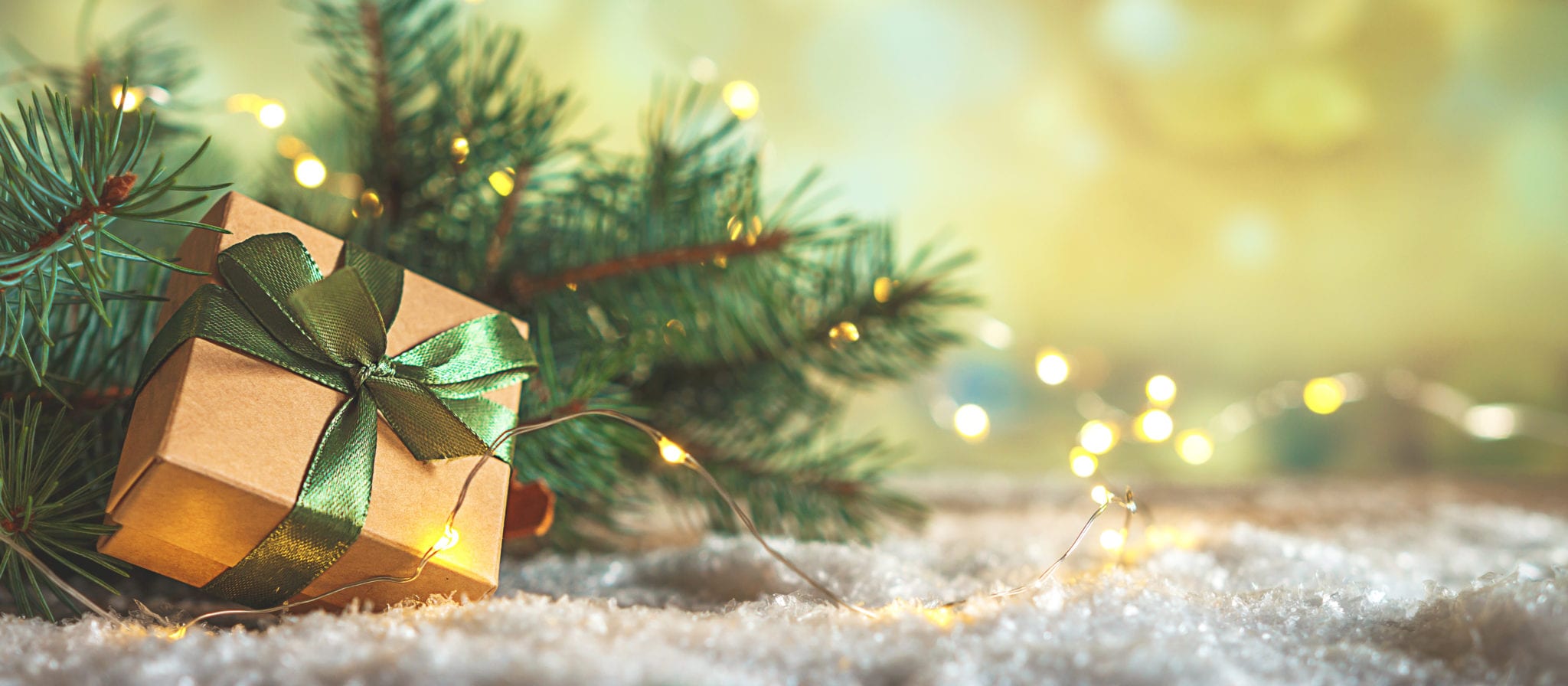 Christmas gift box with green ribbon next to pine branches and festive lights