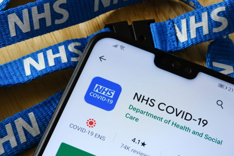 NHS COVID-19 app seen in Play Store on the screen of smartphone next to NHS lanyard.
