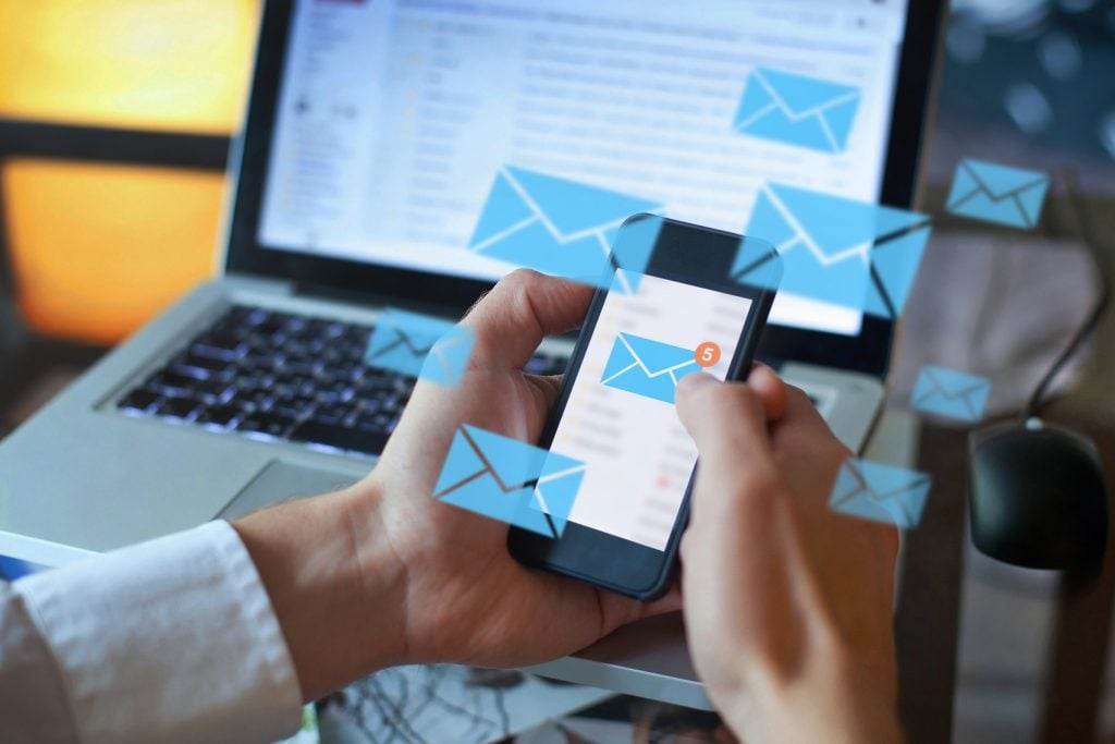 Person checking email on a smartphone with email icons floating over a laptop screen in the background