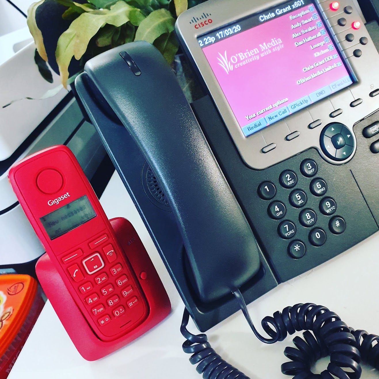 Red cordless telephone handset on a white desk next to a blueish grey desk office phone with a pink screen.